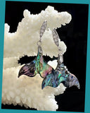 A pair of abalone shell mermaid tail earrings with cubic zirconia embellishments and sterling silver hooks, displayed against a rocky background.