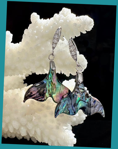 A pair of abalone shell mermaid tail earrings with cubic zirconia embellishments and sterling silver hooks, displayed against a rocky background.