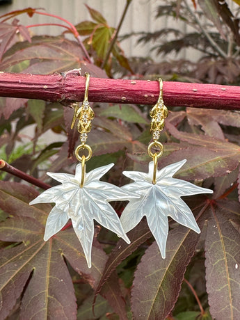 A pair of maple leaf-shaped earrings with gold-plated hooks and cubic zirconia embellishments, displayed against a backdrop of red maple leaves.